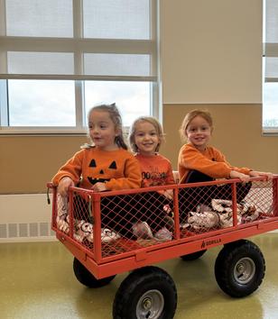 Three children in an orange wagon, wearing Halloween-themed sweaters, smiling.