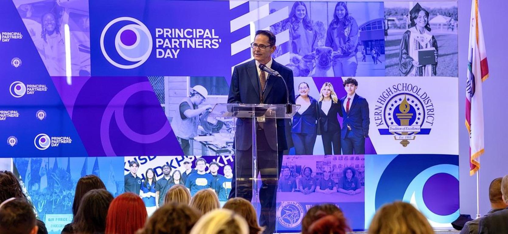 Man speaking at a podium during Principal Partners Day event with audience.