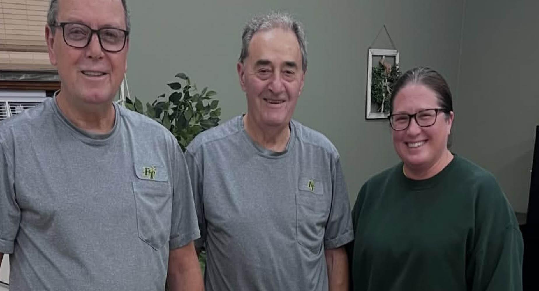 Three adults stand together, wearing gray shirts, in a warm, indoor setting.