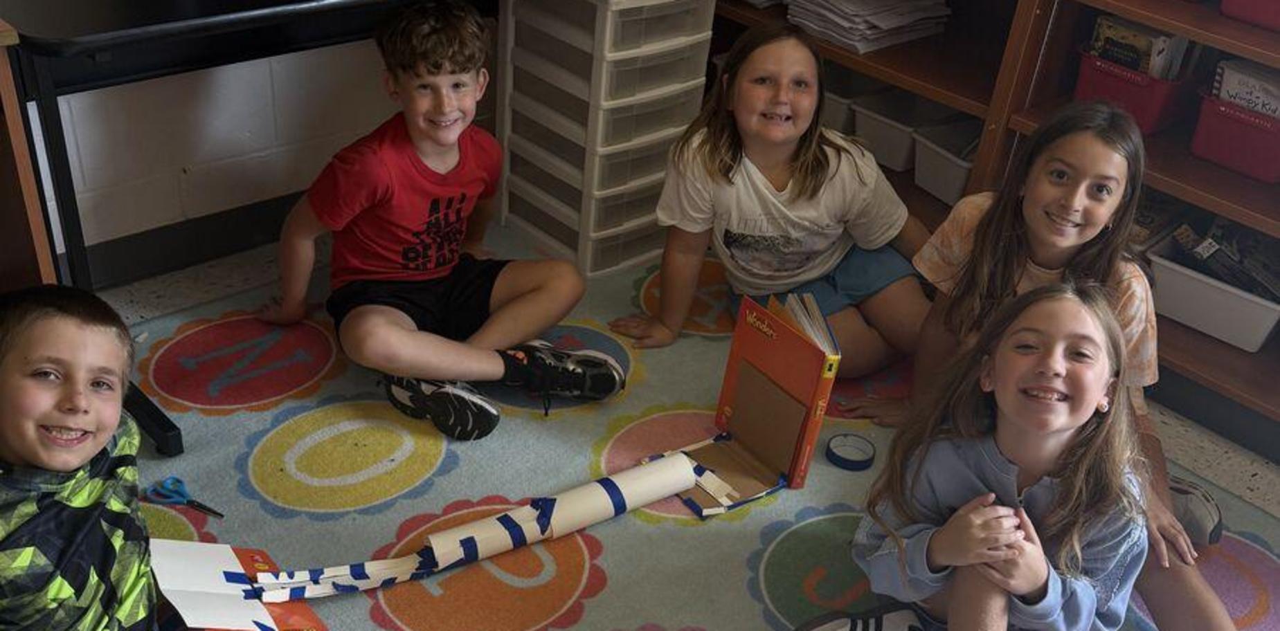 Five children sitting on a colorful rug, smiling around a homemade project.