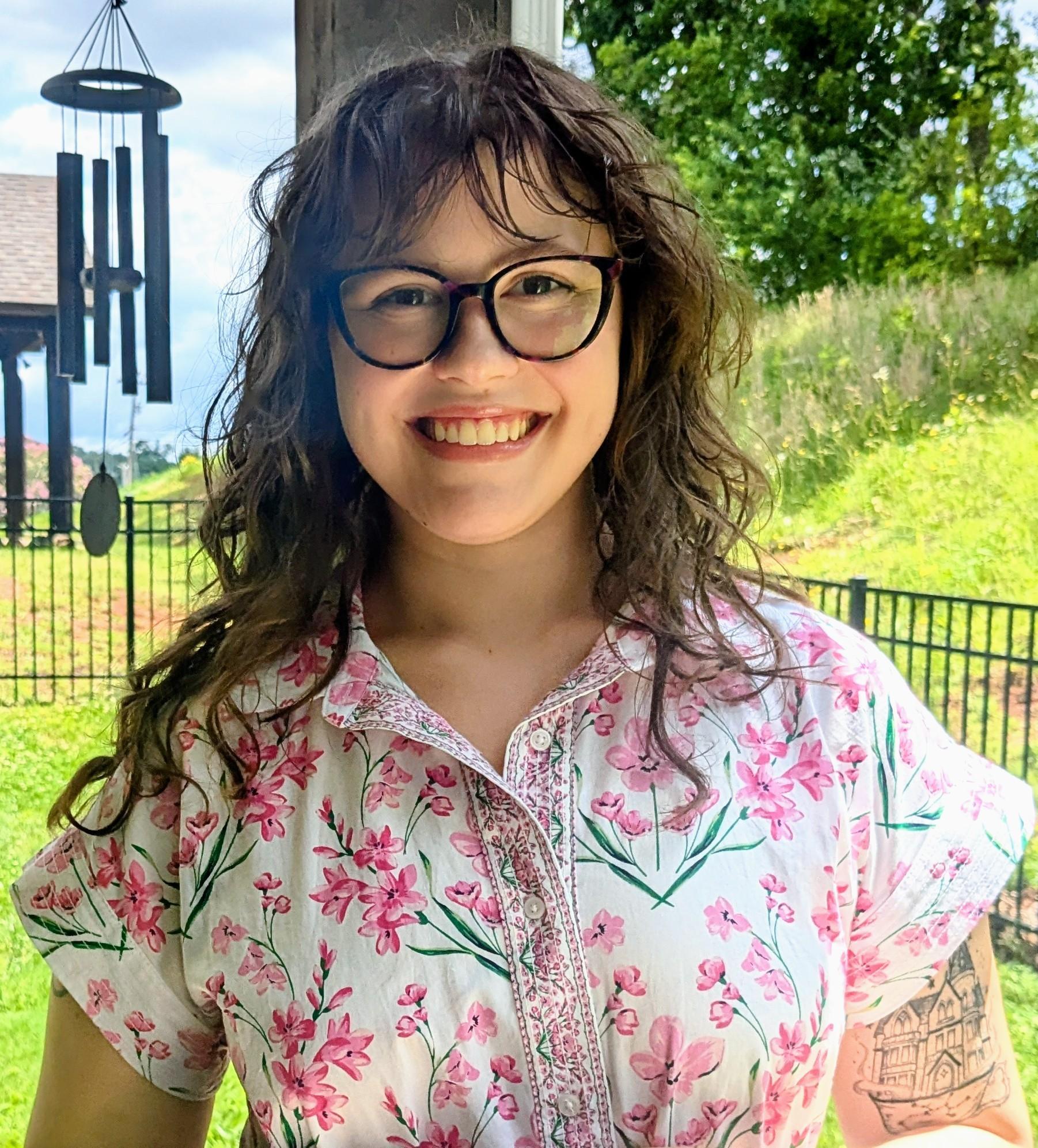 Smiling person with glasses wearing a floral shirt in a garden setting.