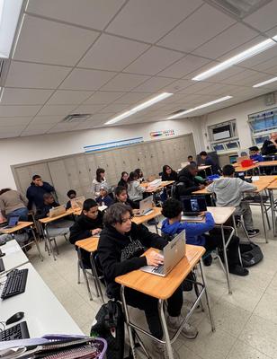 Students working on laptops in a classroom with desks and chairs.