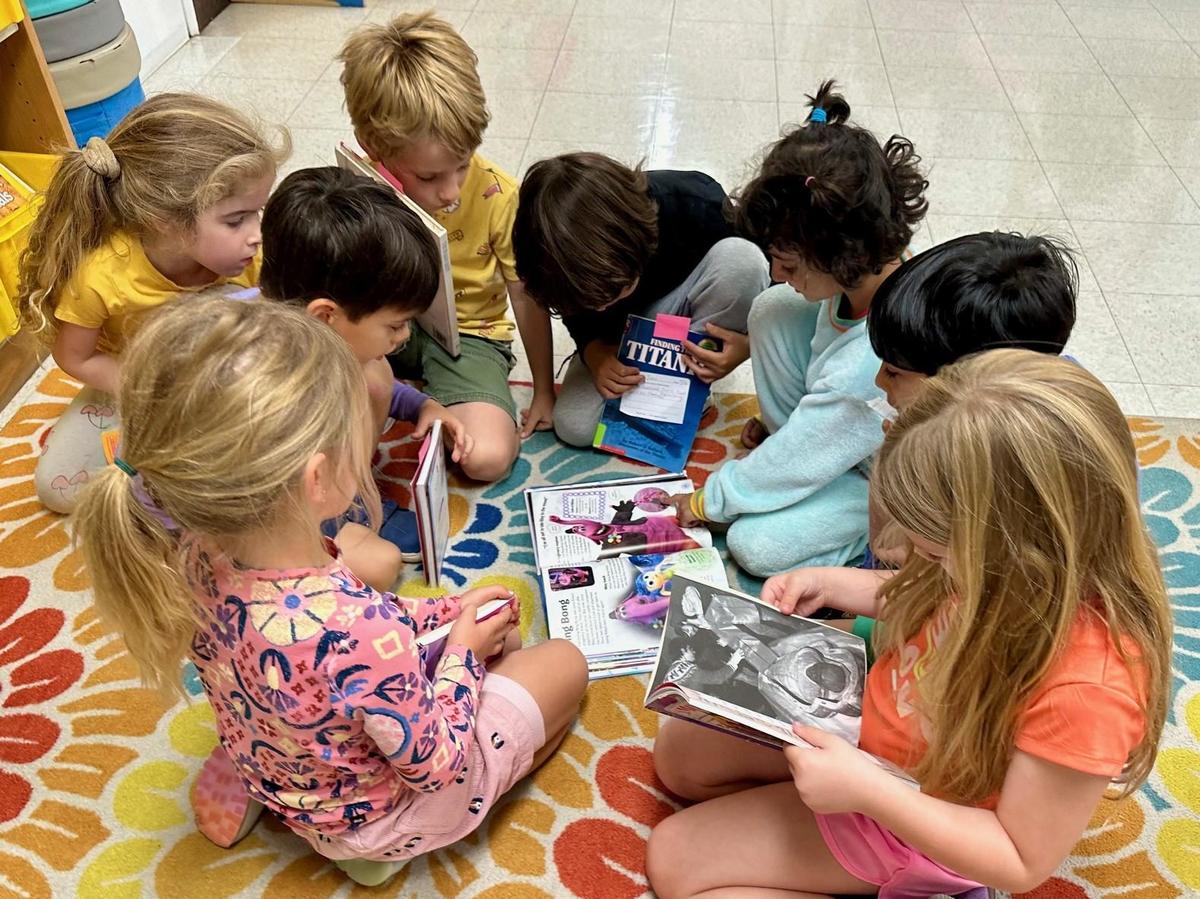 Kindergarten students gather on a rug, looking at books together.
