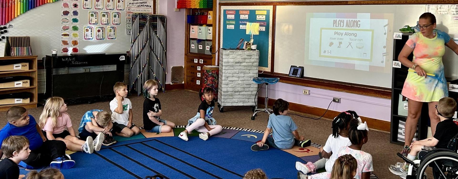 Children sitting on a carpet attending a lesson from a teacher with a presentation.