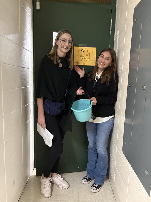 Two girls in costumes stand in a hallway holding a bucket and smiling.