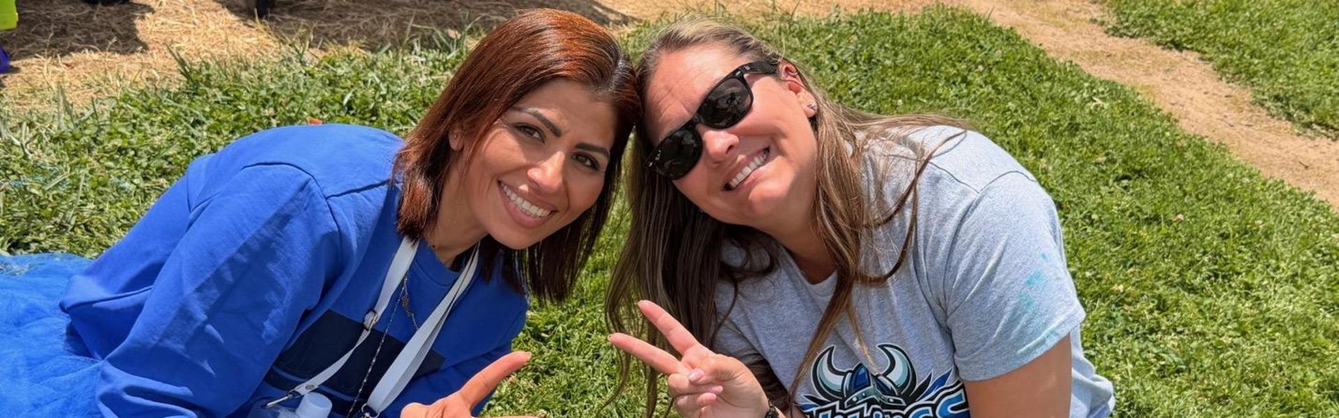Two smiling women in casual clothing making peace signs on green grass.