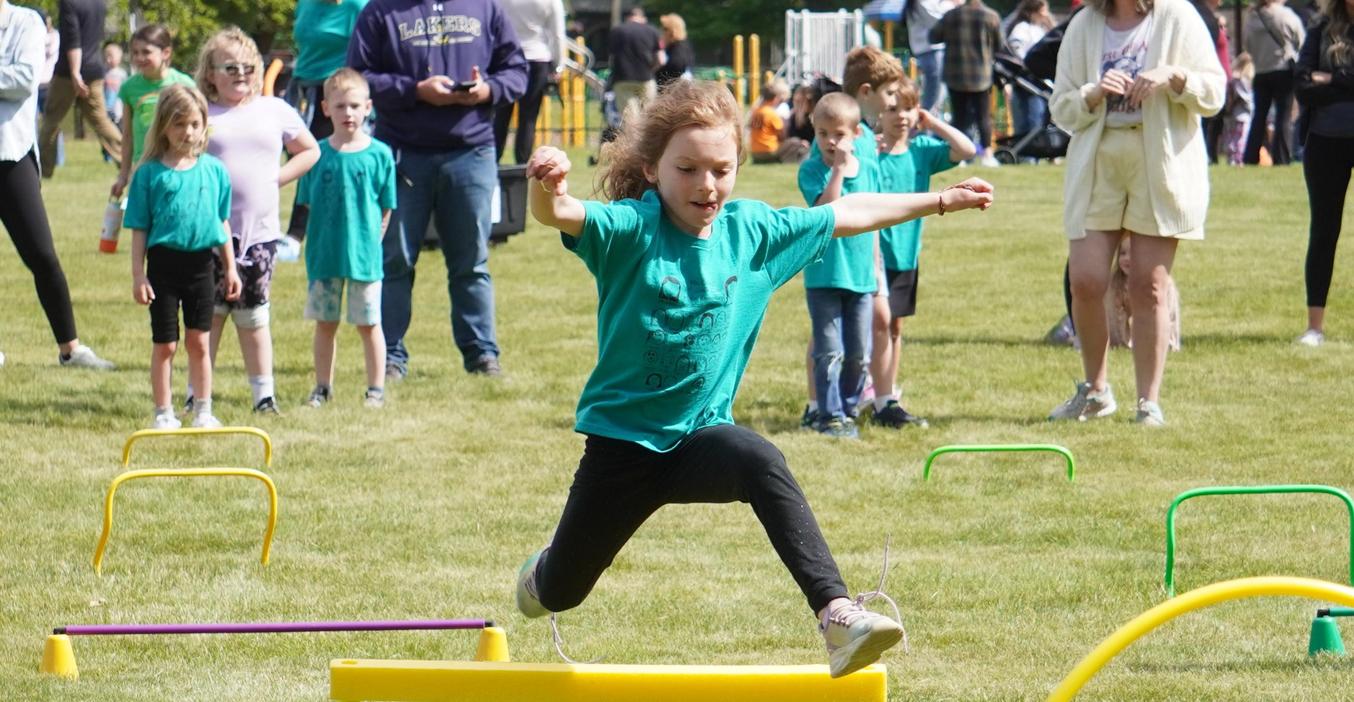 A child in a green shirt leaps over a low hurdle during an outdoor event.