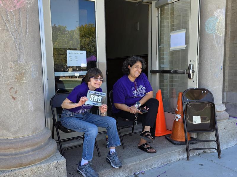 Two women in purple shirts sitting on chairs outside a building, one holding a sign.