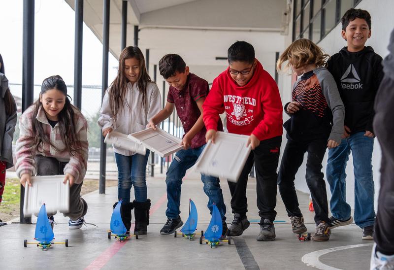 Elementary students line up outside, racing their LEGO-built sail cars by using plastic bin lids to create wind. The students are smiling and fully engaged, showing teamwork and excitement during the STEAM activity.