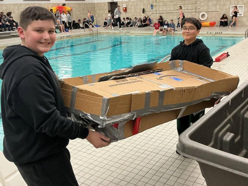 Two boys carrying a large, taped cardboard piece near a swimming pool.