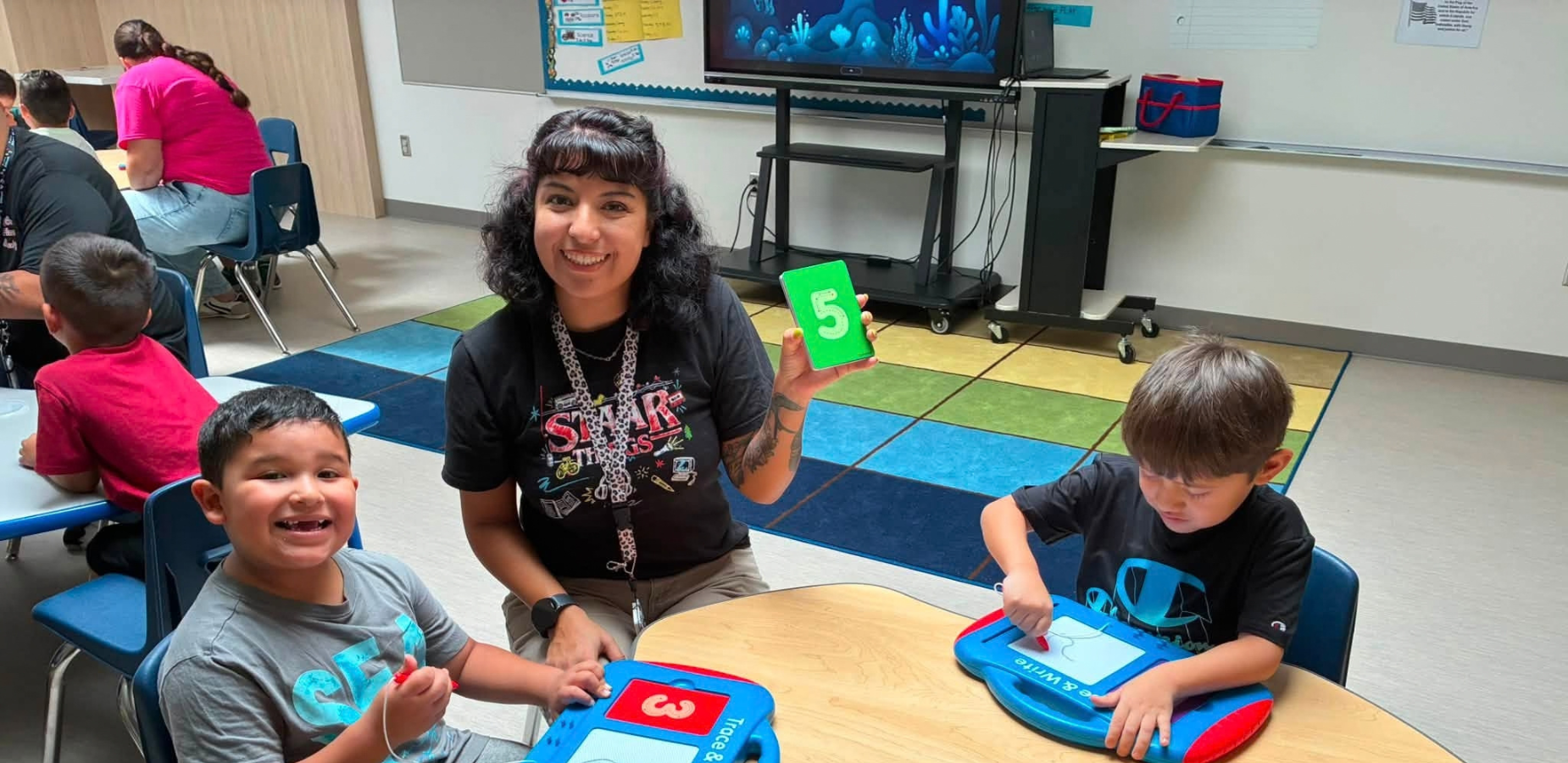 A teacher with two children showing number cards in a classroom setting.