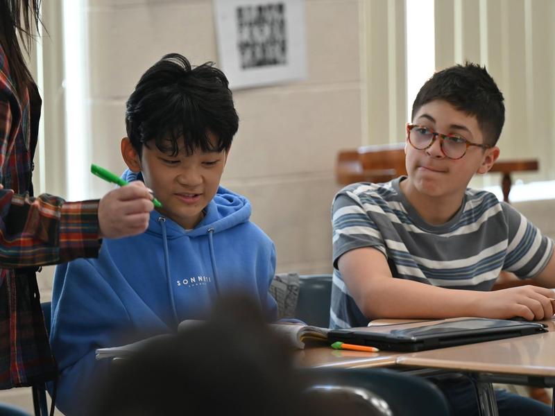 Two students focus on their tasks at a classroom desk with a teacher.