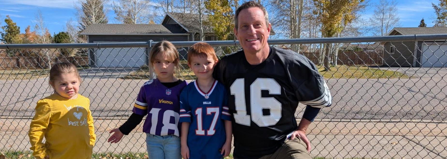 Three children in NFL jerseys pose with an adult male in a sports shirt outdoors.