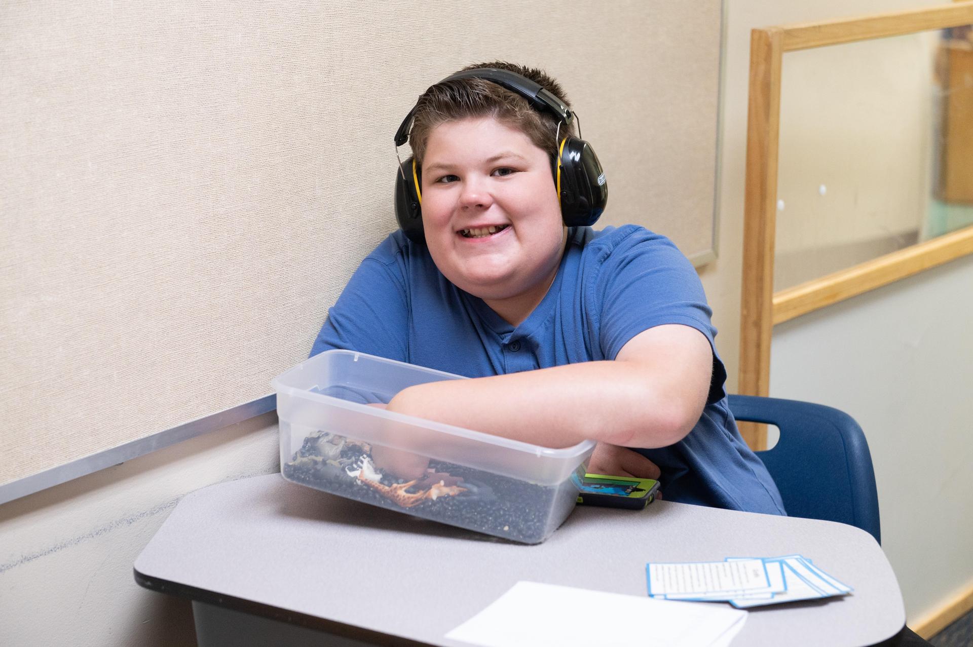 Student smiling at camera wearing blue shirt sitting at desk wearing headphones and choosing crayon.