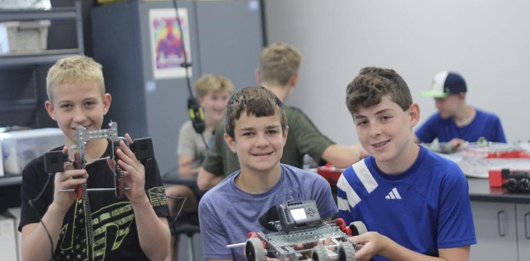 Three boys proudly displaying their completed robot in a classroom.