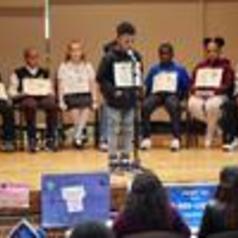 A group of students sitting on a stage, with one boy speaking into a microphone.