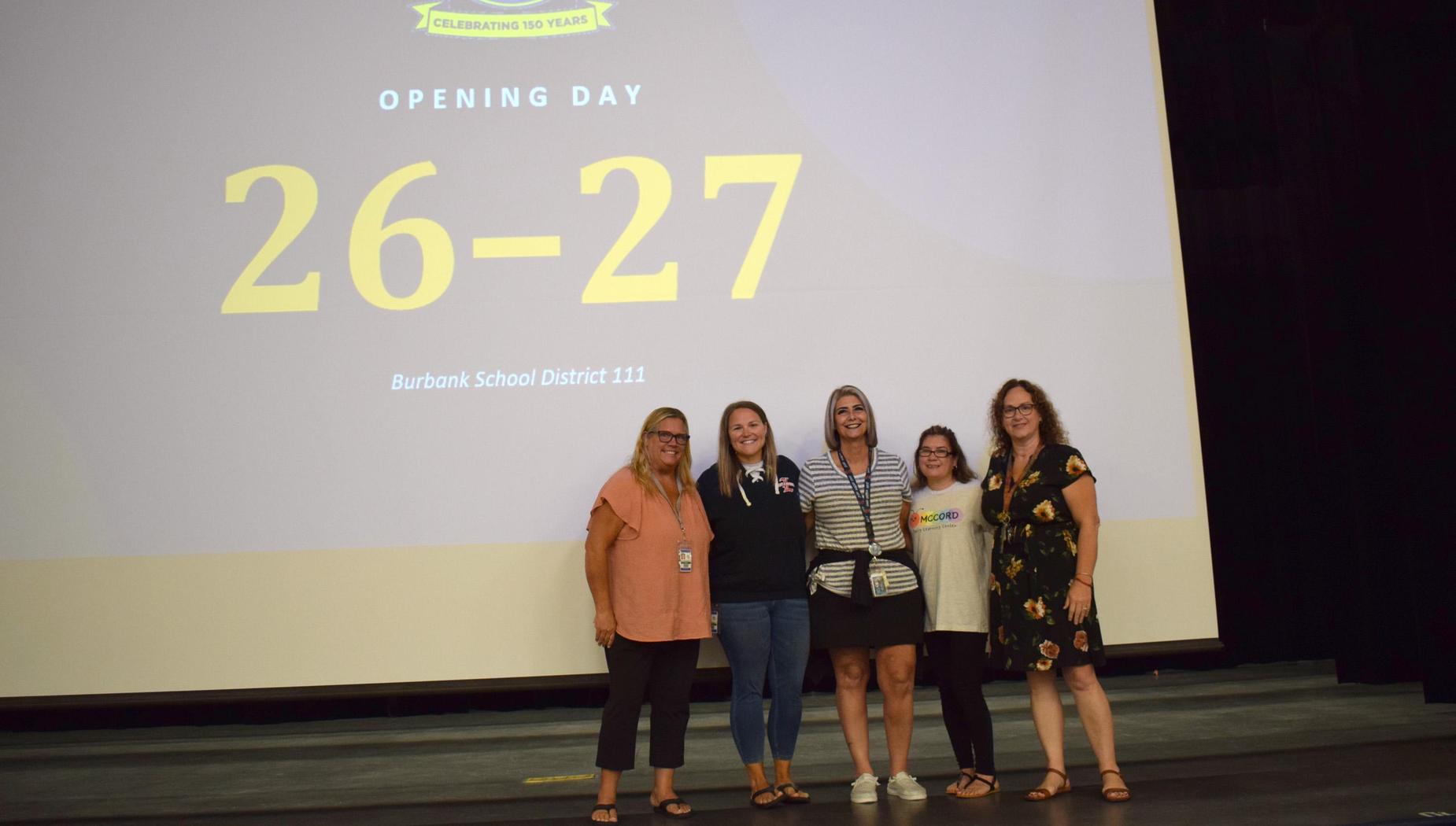 Five individuals posing on stage in front of a screen displaying event details.