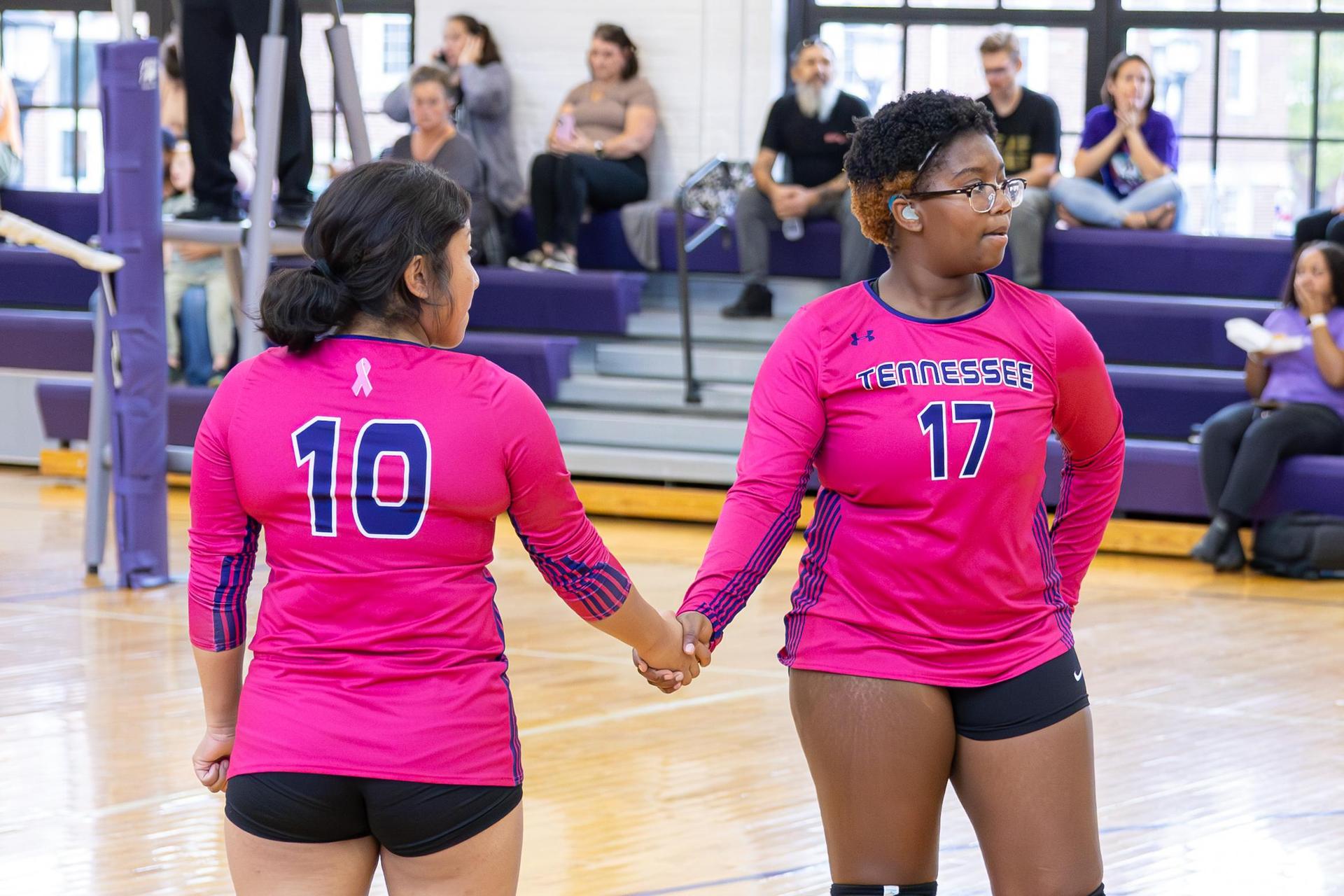 Two volleyball players, one in pink and the other in black, hold hands before a match.