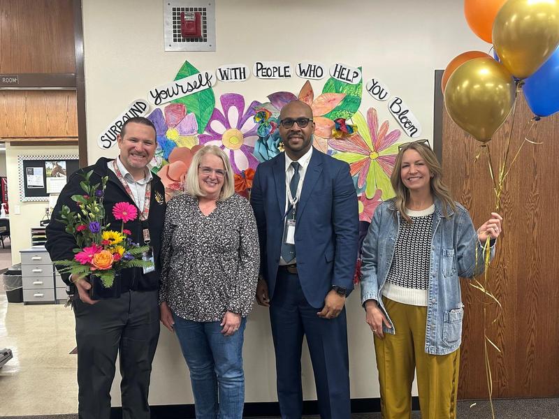 Four adults pose together in front of a colorful flower mural, holding balloons and a bouquet.