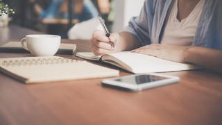 Close-up of hands writing in a notebook at a table with a cup and phone