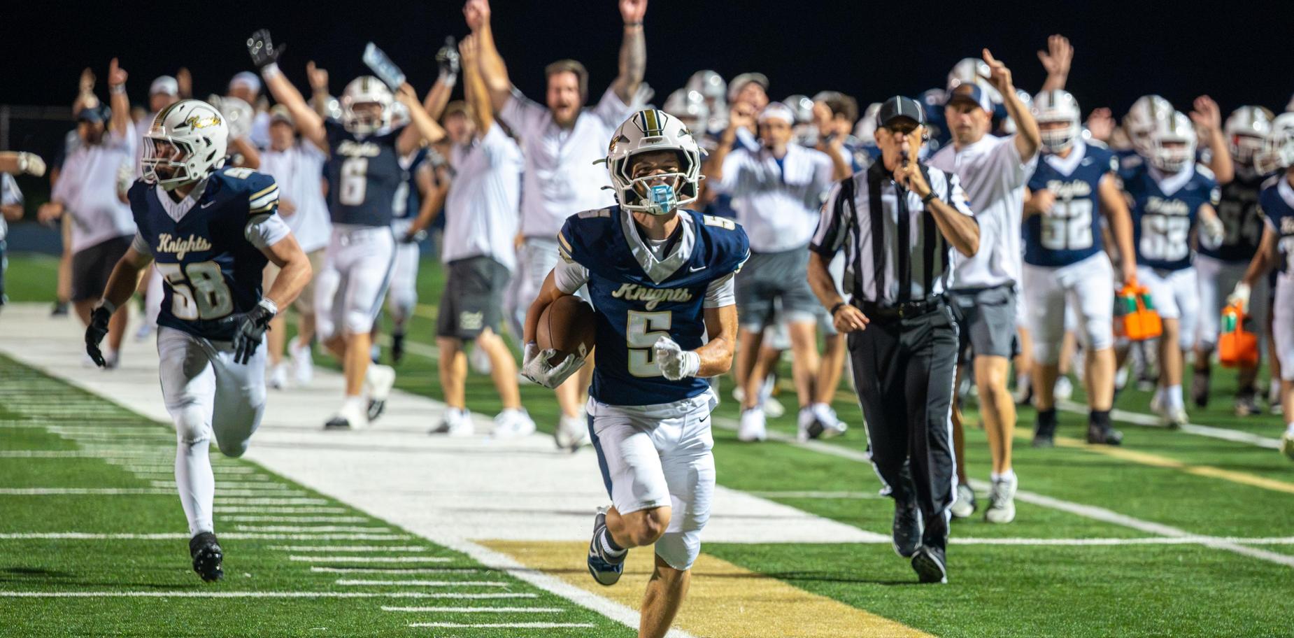 A football player runs towards the end zone with teammates and fans cheering in the background.
