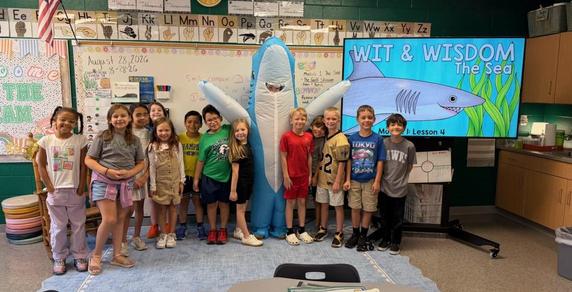 Children in a classroom pose with a large shark costume and a screen displaying ocean-themed content.