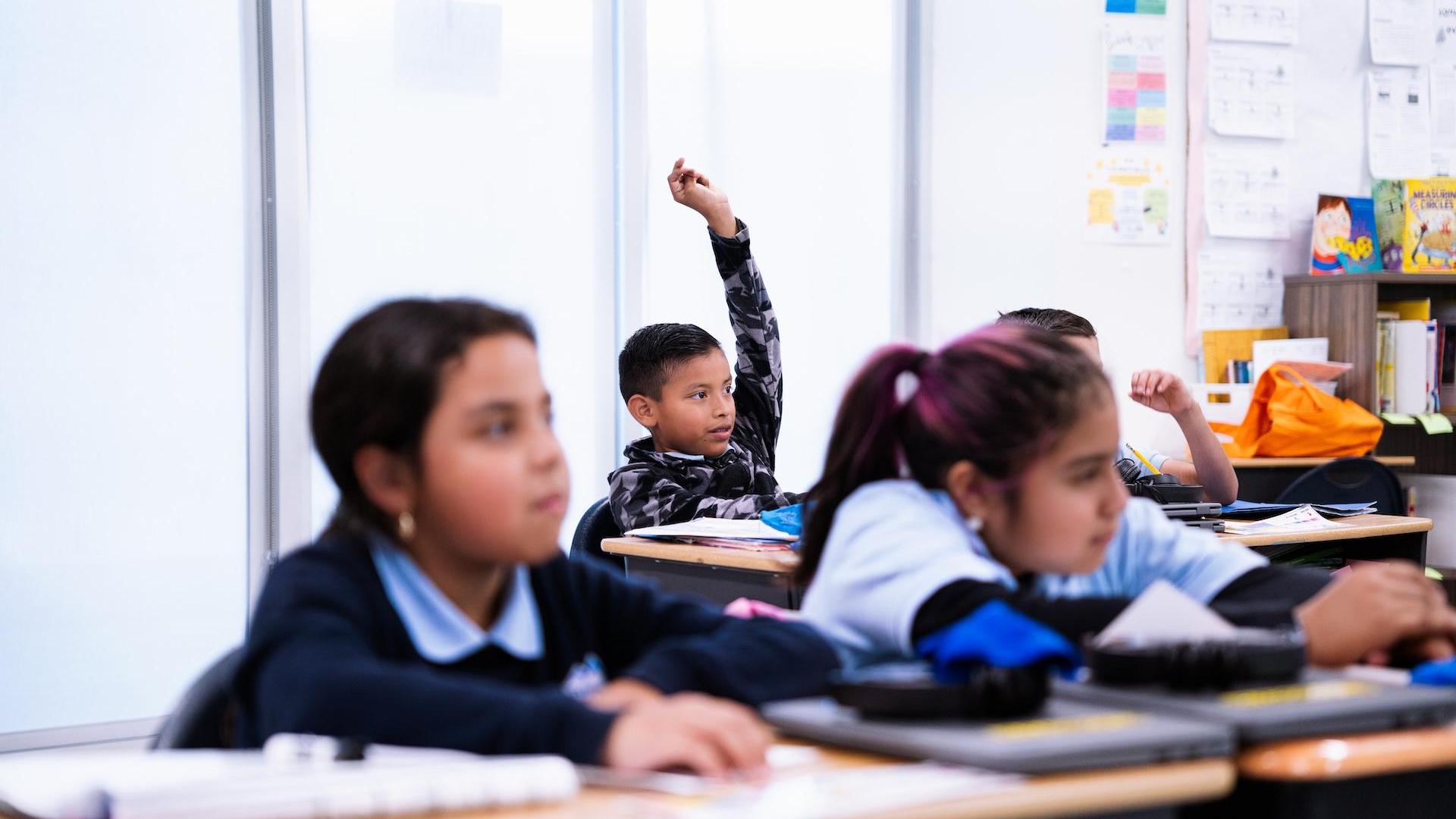 Group of children in a classroom, one boy raising his hand to speak.