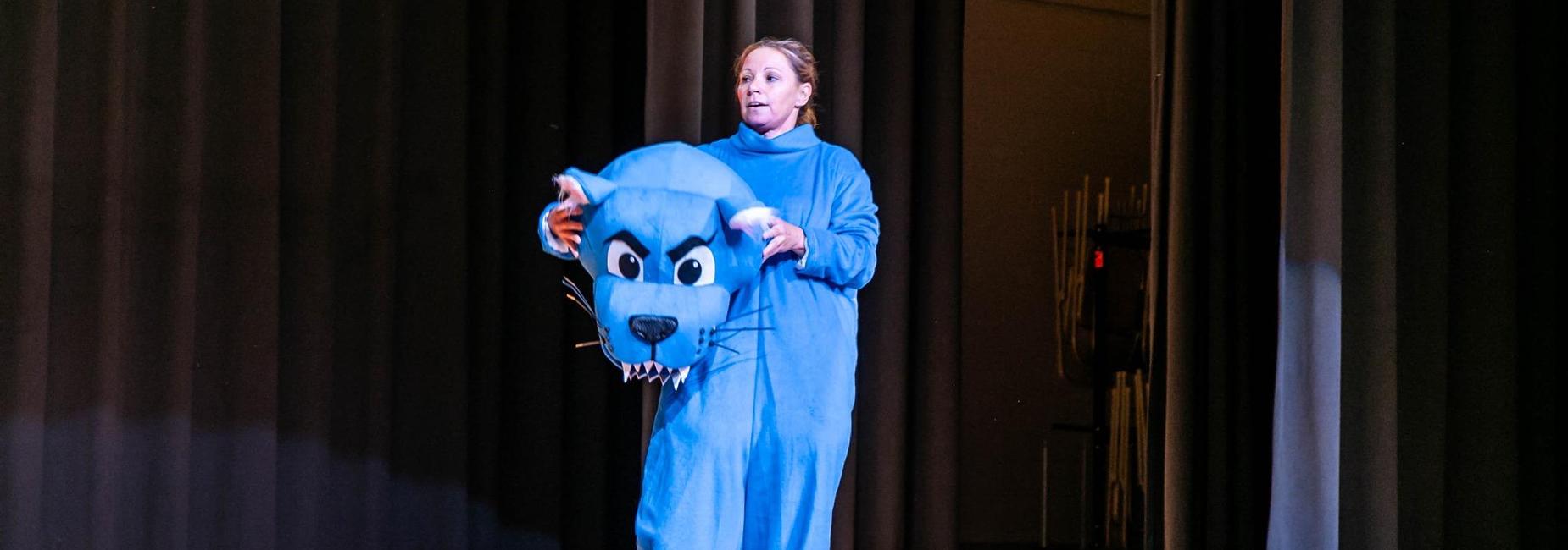 Woman wearing a large blue cat costume on stage with curtains in the background.