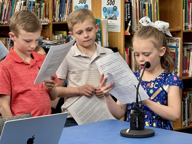 A young girl reads and speaks into a mic as two other students listen