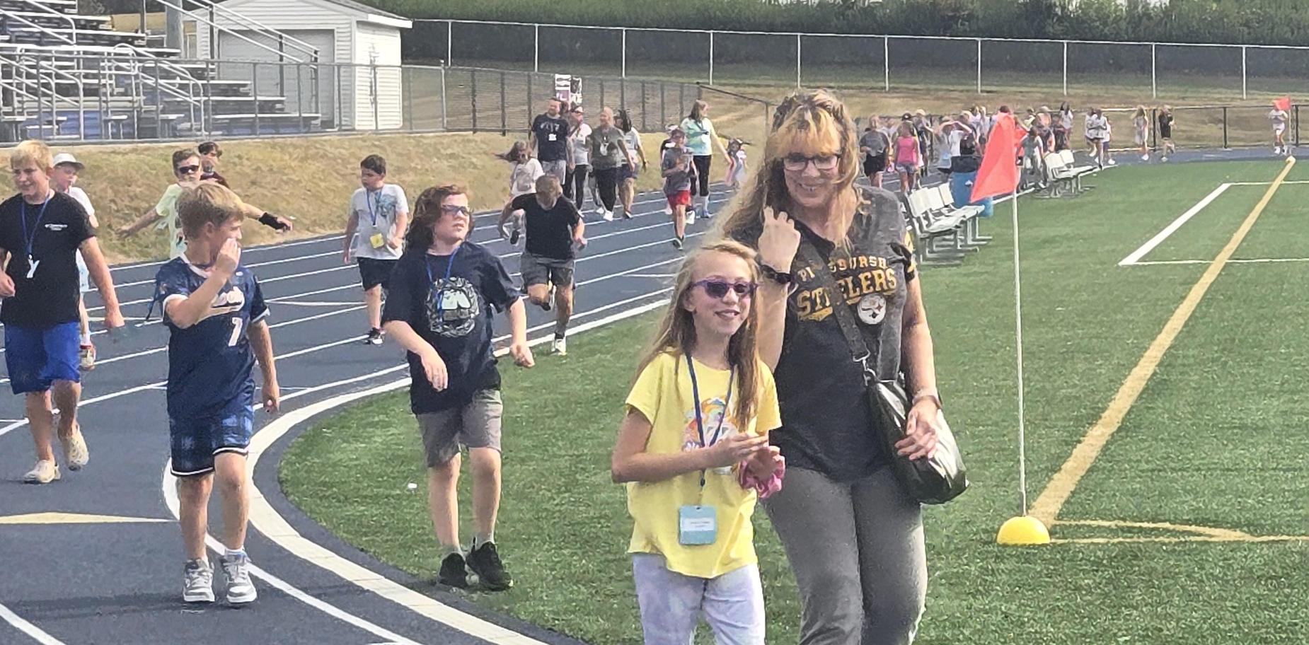 Children walking on a track, with an adult and a sports facility in the background.
