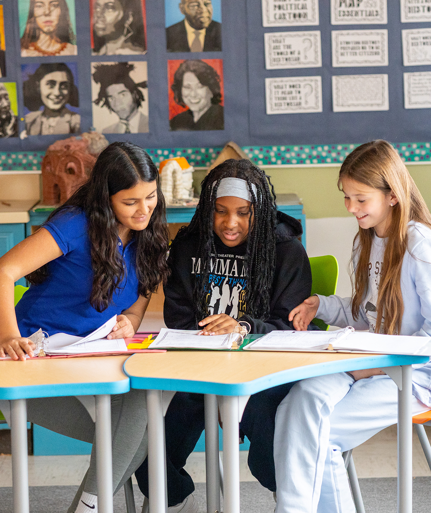 Three students sitting together at connected desks, collaborating on an assignment using open binders in a classroom.