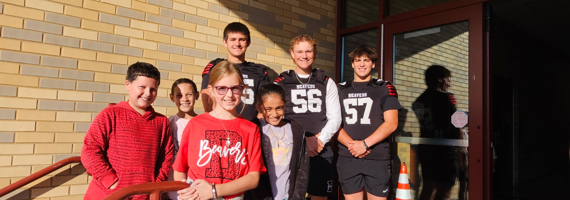 Group of younger children with football players, all smiling together by the school entrance.
