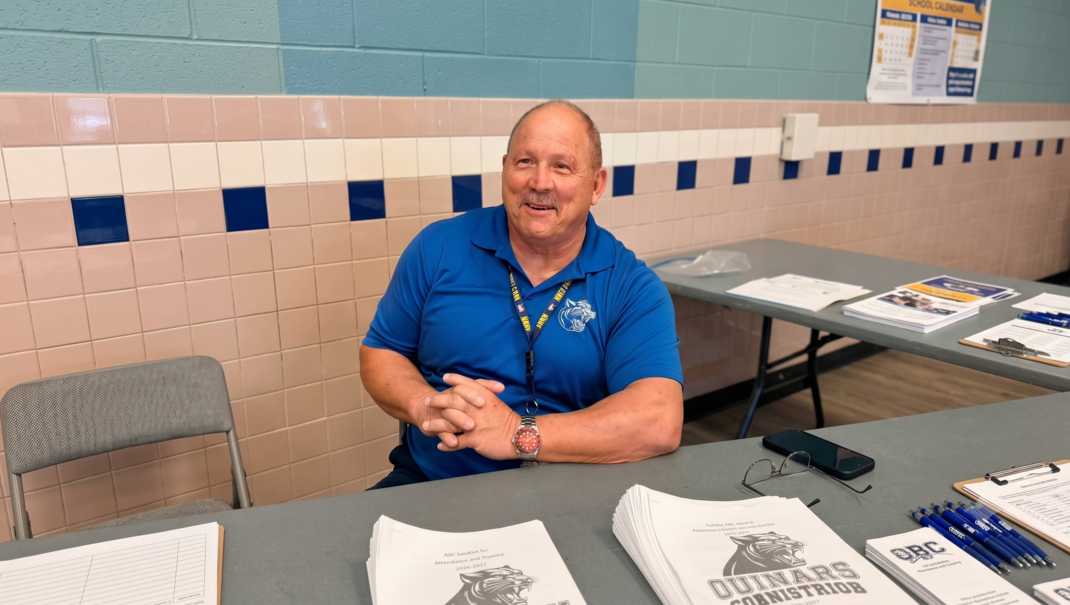 A smiling man in a blue shirt sitting at a table with documents and a phone.