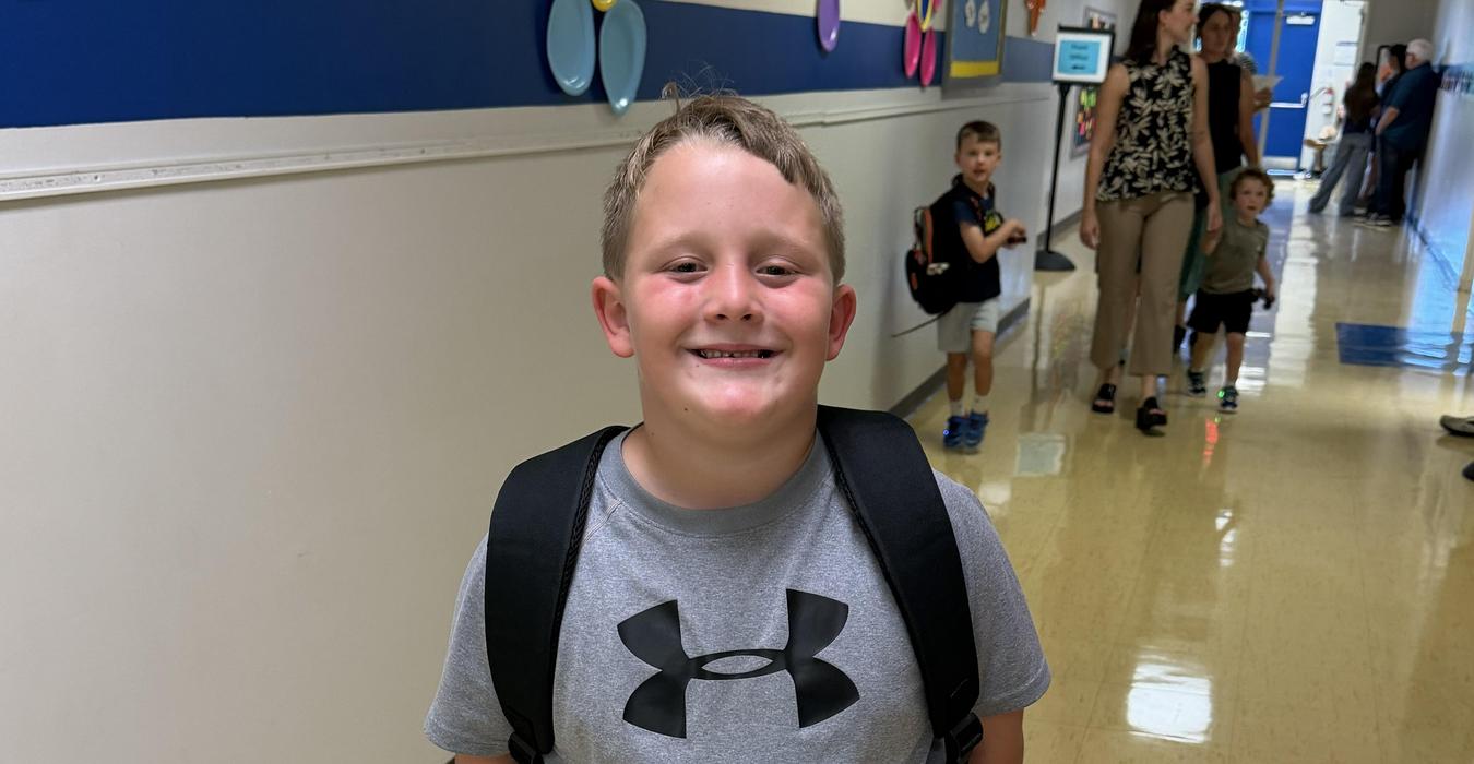 Smiling boy in a gray shirt with a backpack in a school hallway.