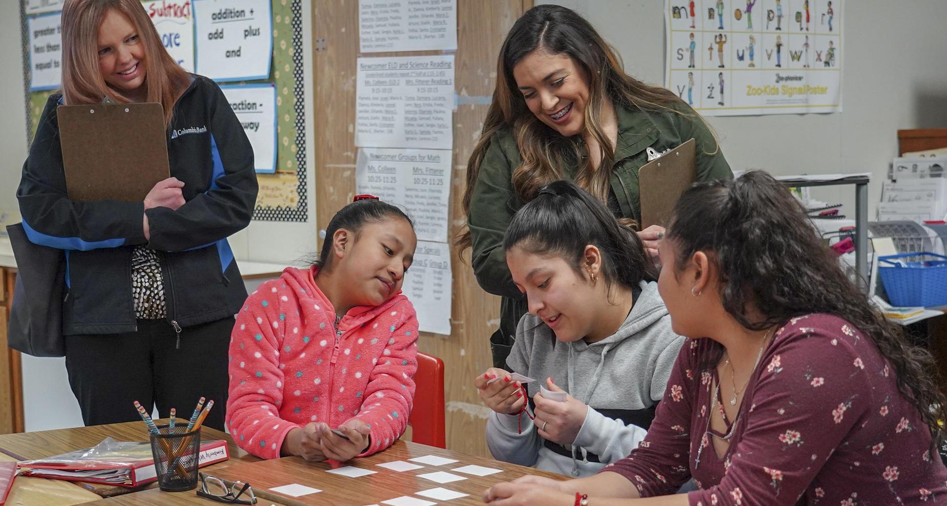 Students looking at flashcards while two female adults are watching them.