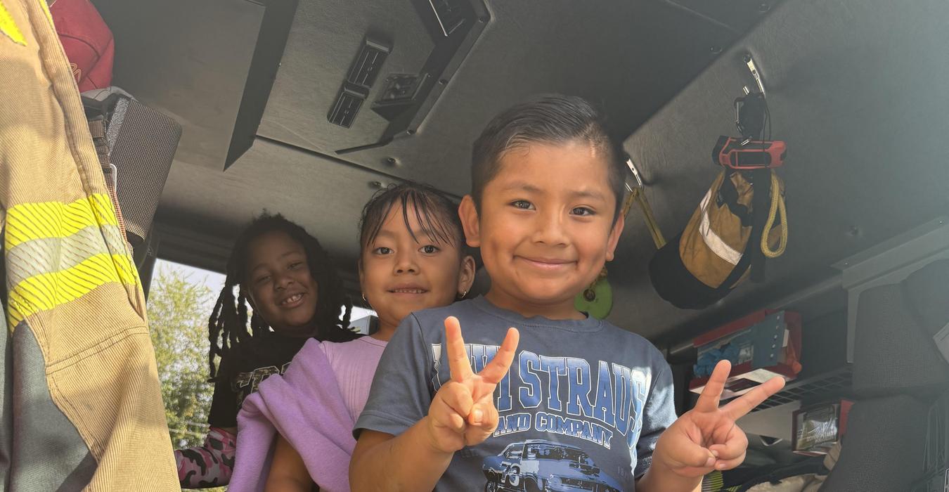 Three smiling kids inside a fire truck, posing cheerfully with peace signs.