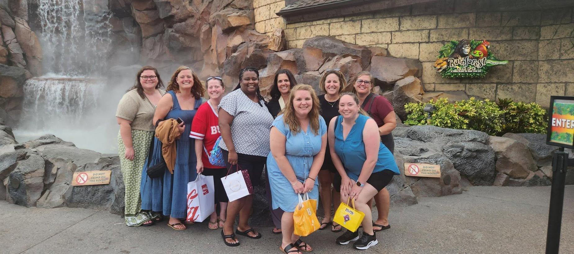A group of nine women posing together in front of a rock formation and a water backdrop.
