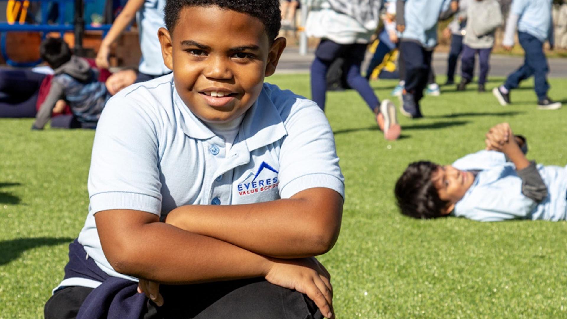 A boy sitting on grass, smiling, with children playing in the background.