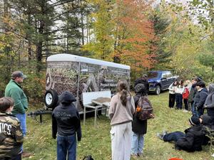 A group of people gathered around a trailer with educational materials in a forested area.
