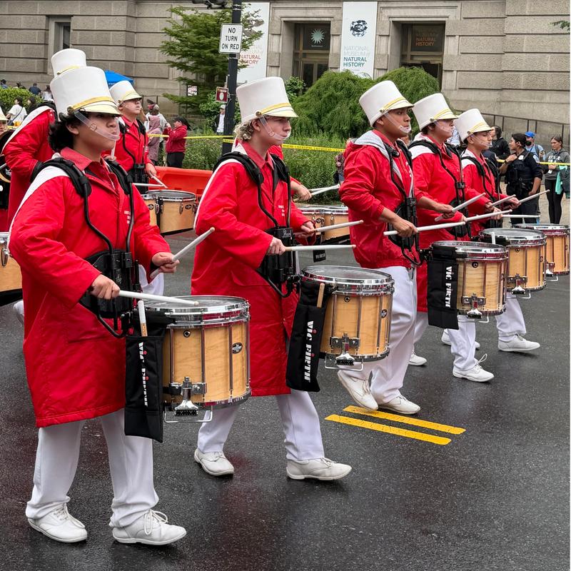 Marching band members in red coats and white hats playing drums in a parade.