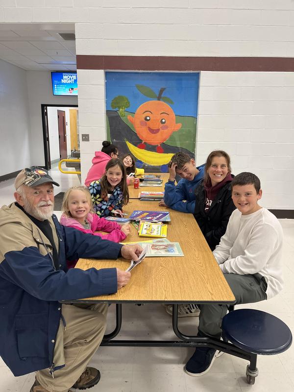 Elderly man and children gathered around a brown table with books in a school cafeteria.