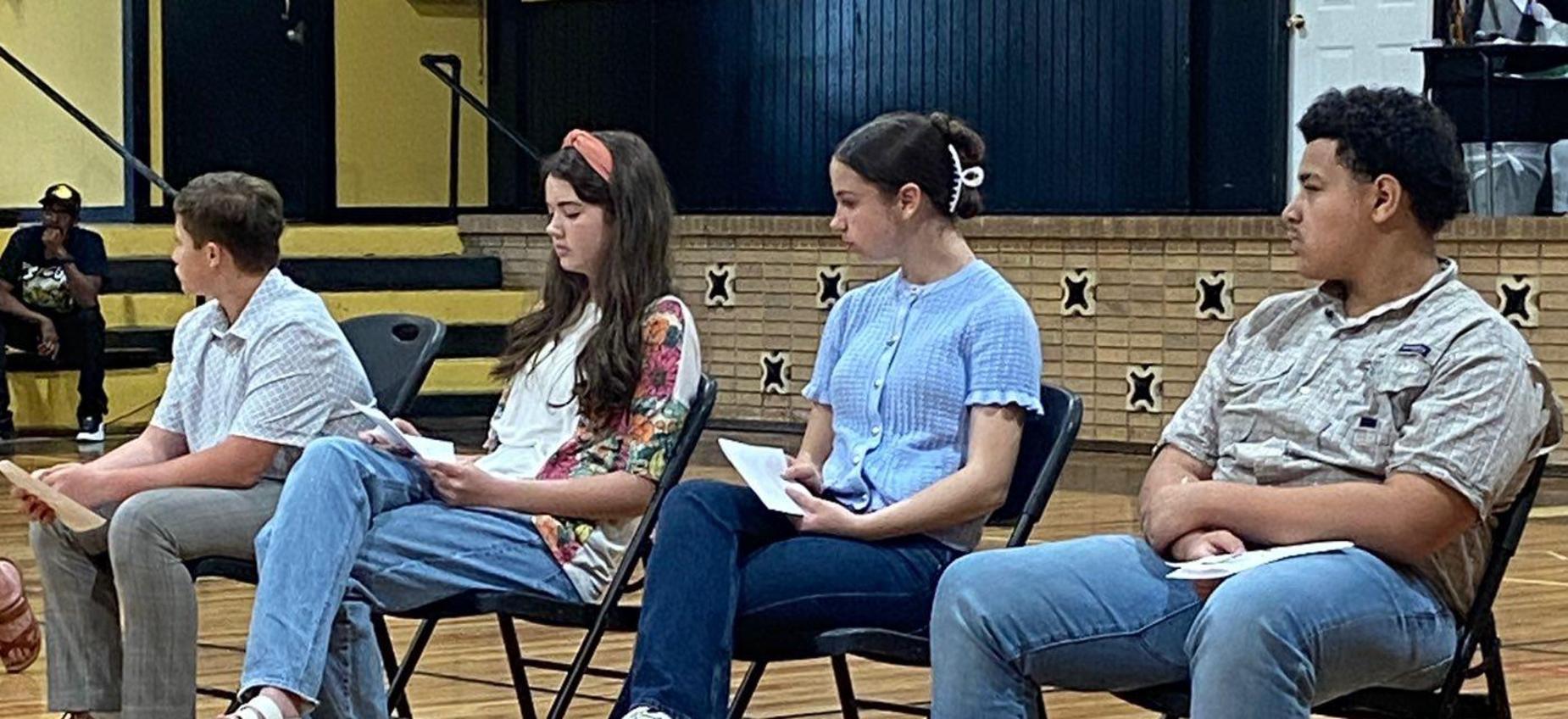 Four individuals sitting on chairs, each holding a paper in a gymnasium.