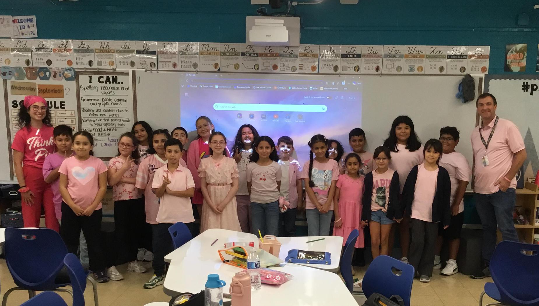 Group of students and adults wearing pink, smiling in a classroom setting.