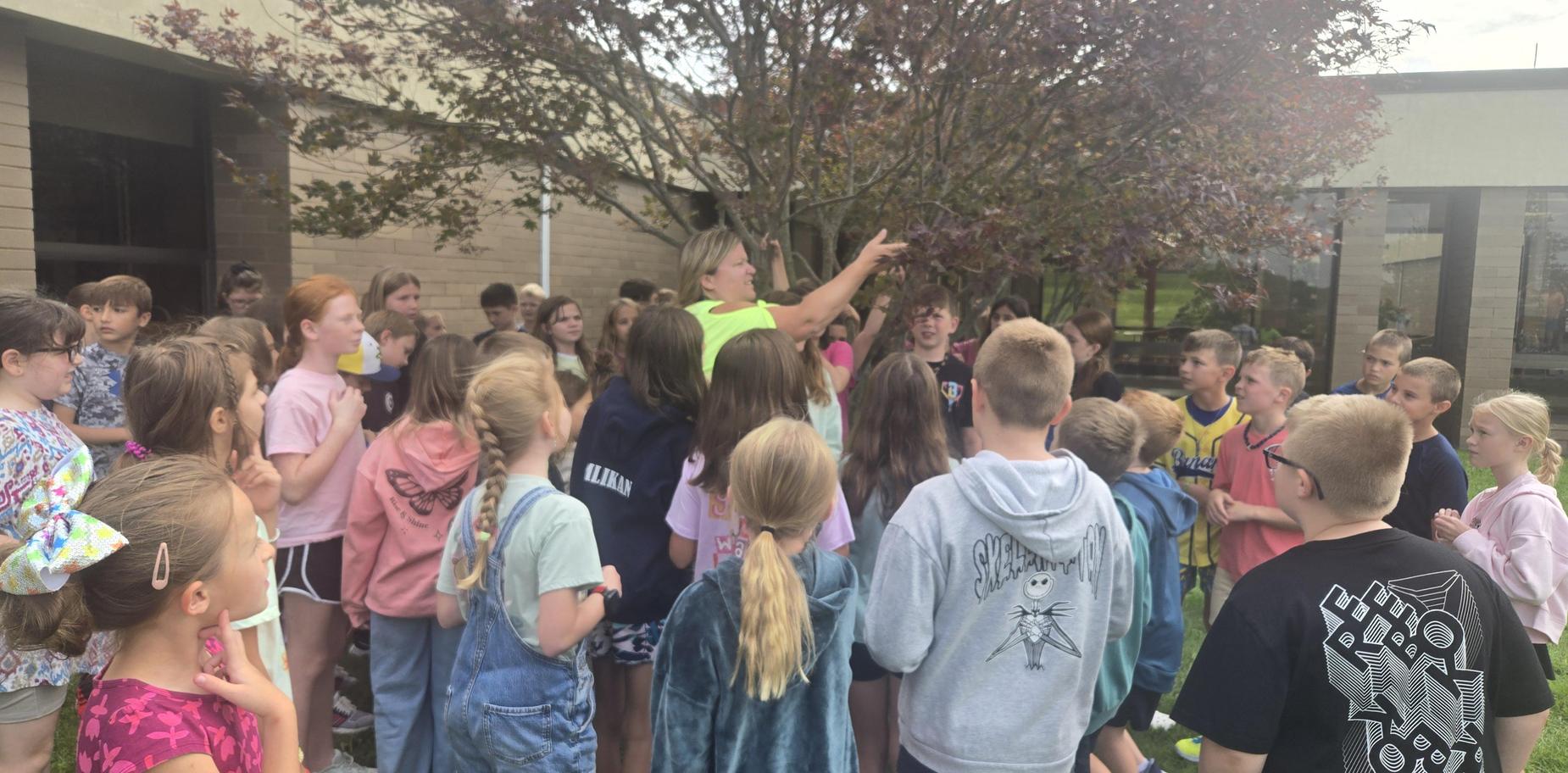 Group of children gathered around an adult under a tree during an outdoor activity.