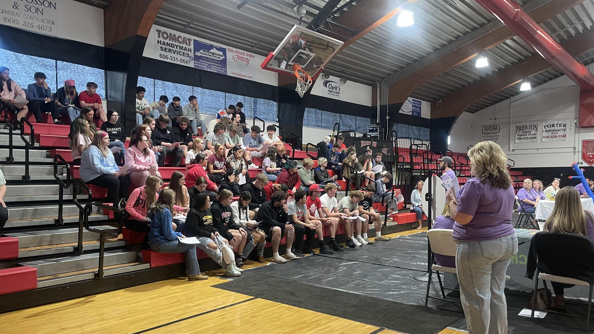 Crowd of students seated in a gym, listening to a speaker at an event.