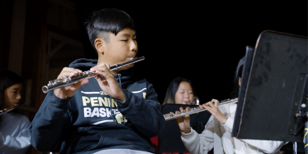 A boy plays the flute on stage with other musicians in the background.