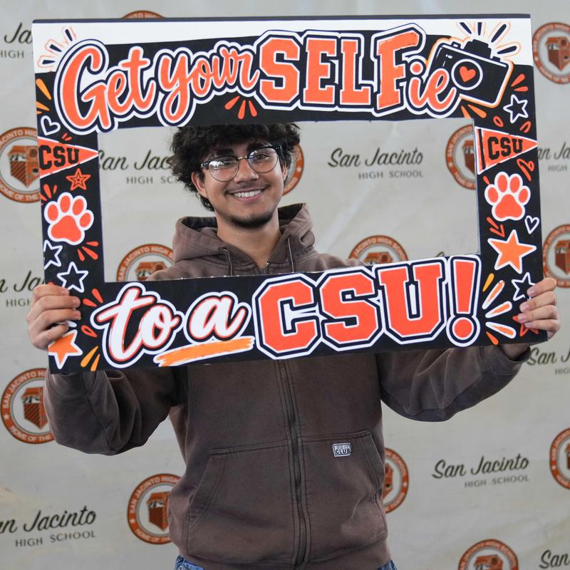 Smiling young man holding a colorful frame that says 'Get your SELFie to a CSU!'