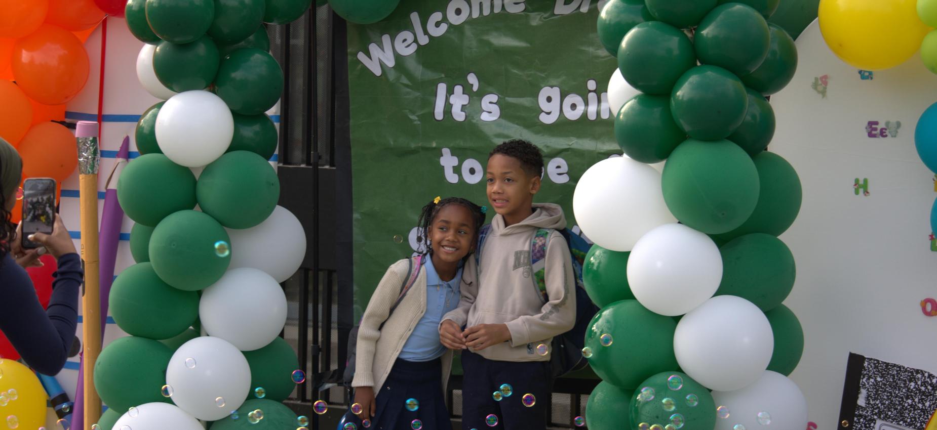 Two children pose together in front of a decorated backdrop with balloons.