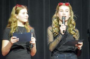 Two girls in black outfits speaking into microphones on stage.