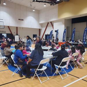 Families with young children gather at tables in a gymnasium during the PUSD Kinder Round-Up event, with school registration booths visible in the background.